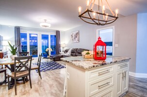 A modern kitchen with a central island, a dining area with chairs and a table, and a view through large windows.