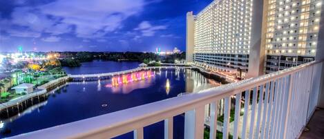 A high-rise hotel with a waterfront view, featuring a well-lit balcony and a reflection of colorful lights on the water.