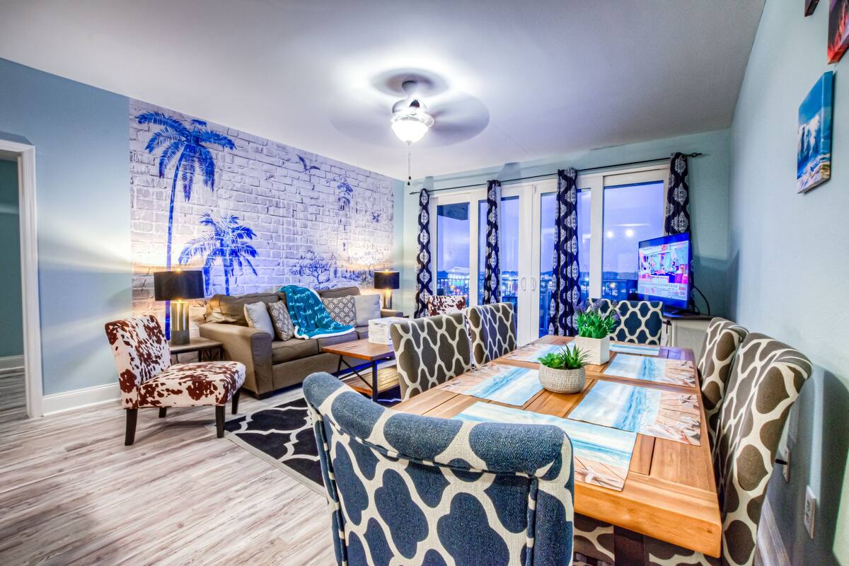 A dining area with a wooden table, patterned chairs, and a brick wall with palm tree artwork.