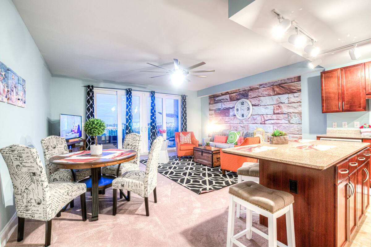 A modern kitchen with a central island, dining area with patterned chairs, and a stone accent wall.