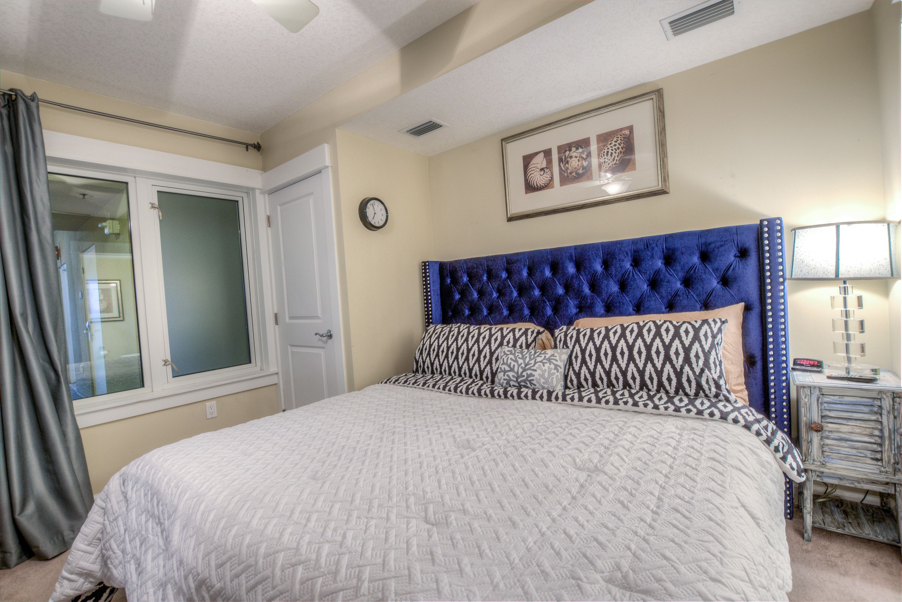 A bedroom with a large bed, a headboard with a tufted design, patterned pillows, a bedside table with a lamp, and a framed picture on the wall.