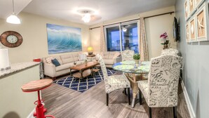 A living room with a large window, a sofa, a coffee table, and a dining area with chairs. - Grand Panama by Aneliya's Rentals (Panama City Beach)