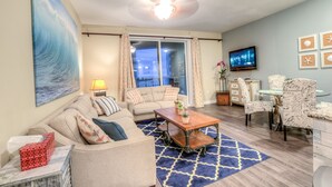 A living room with a sofa, coffee table, and chairs, a TV, and a large window with curtains. - Grand Panama by Aneliya's Rentals (Panama City Beach)