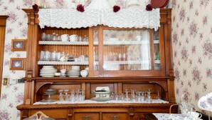 A wooden china cabinet with glassware and dishes, a lace tablecloth, and floral wallpaper. - Garden Gate BnB (Sturgeon Bay)