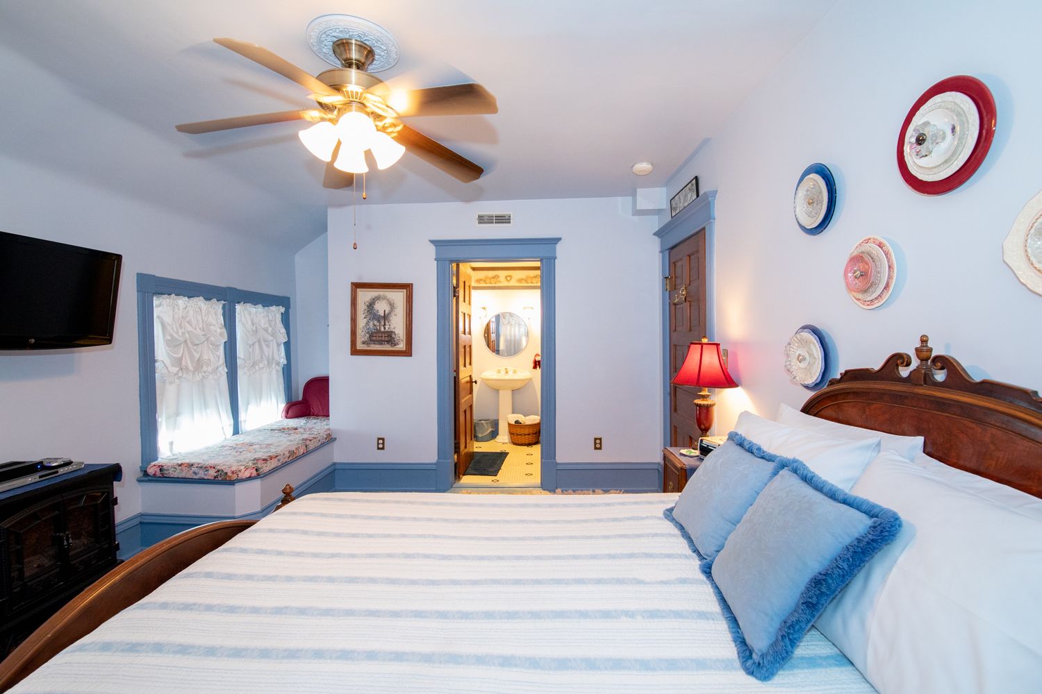 A bedroom with a large bed, a ceiling fan, a television, and a door leading to a bathroom.