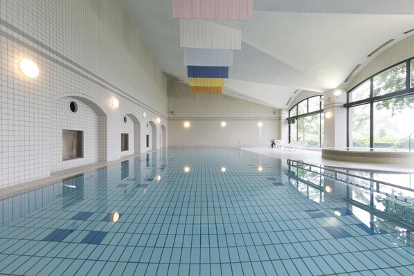 Una piscina interior con paredes y suelo de baldosas, ventanas amplias y un techo alto.