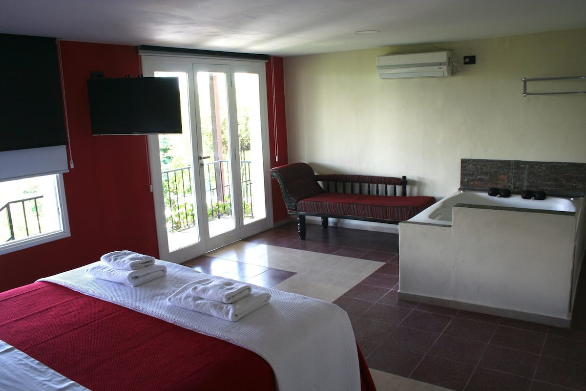 Quarto de hotel com cama, uma cadeira, pequena cozinha e porta de correr para a varanda.