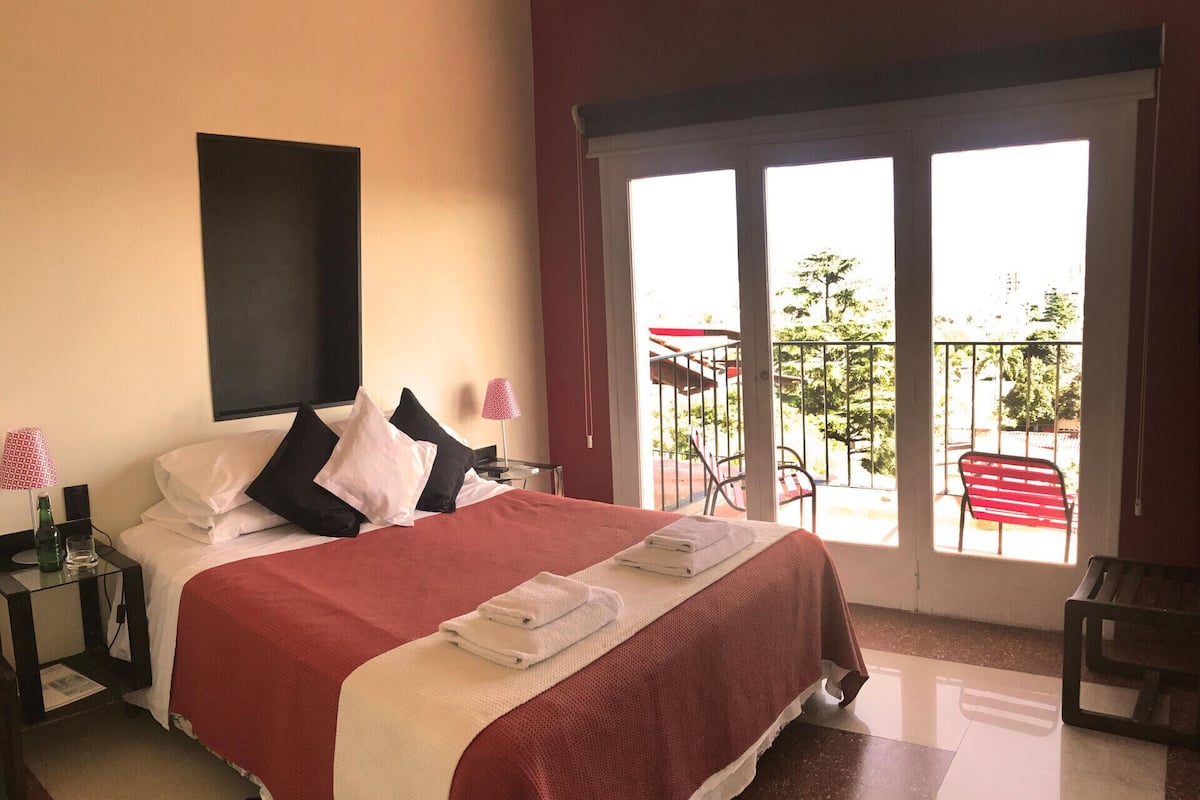 Quarto de hotel com uma cama grande, uma mesa de cabeceira, uma luminária e vista para a varanda externa.
