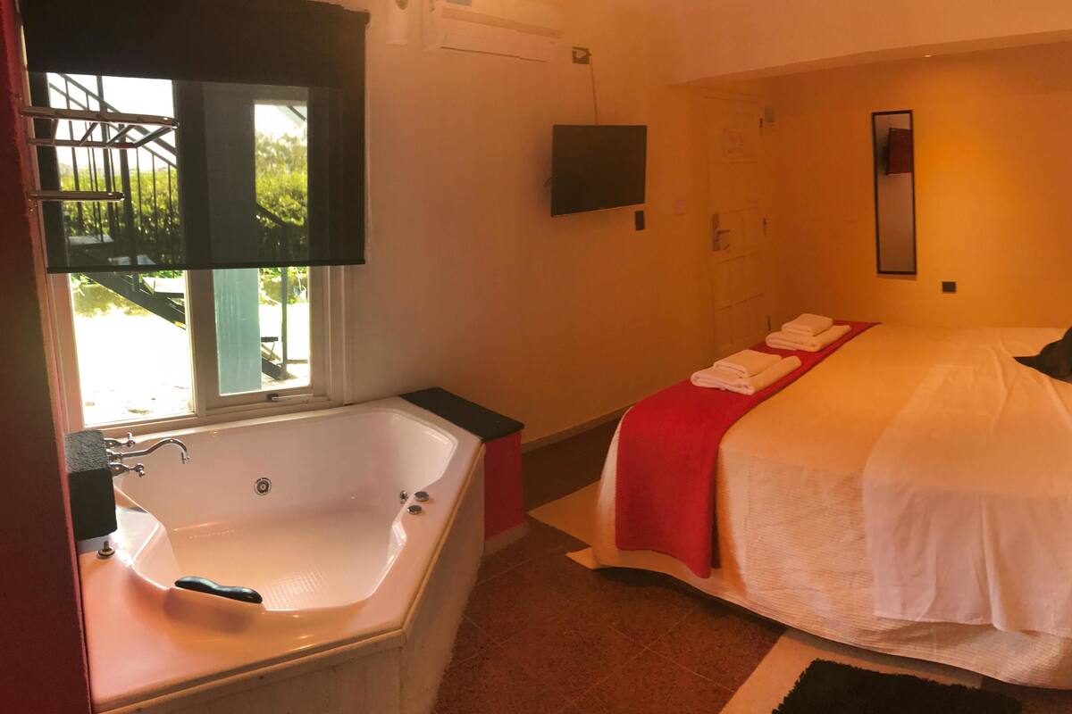 Quarto de hotel com uma banheira grande, cama com lençóis brancos e um detalhe vermelho no cobre-leito.