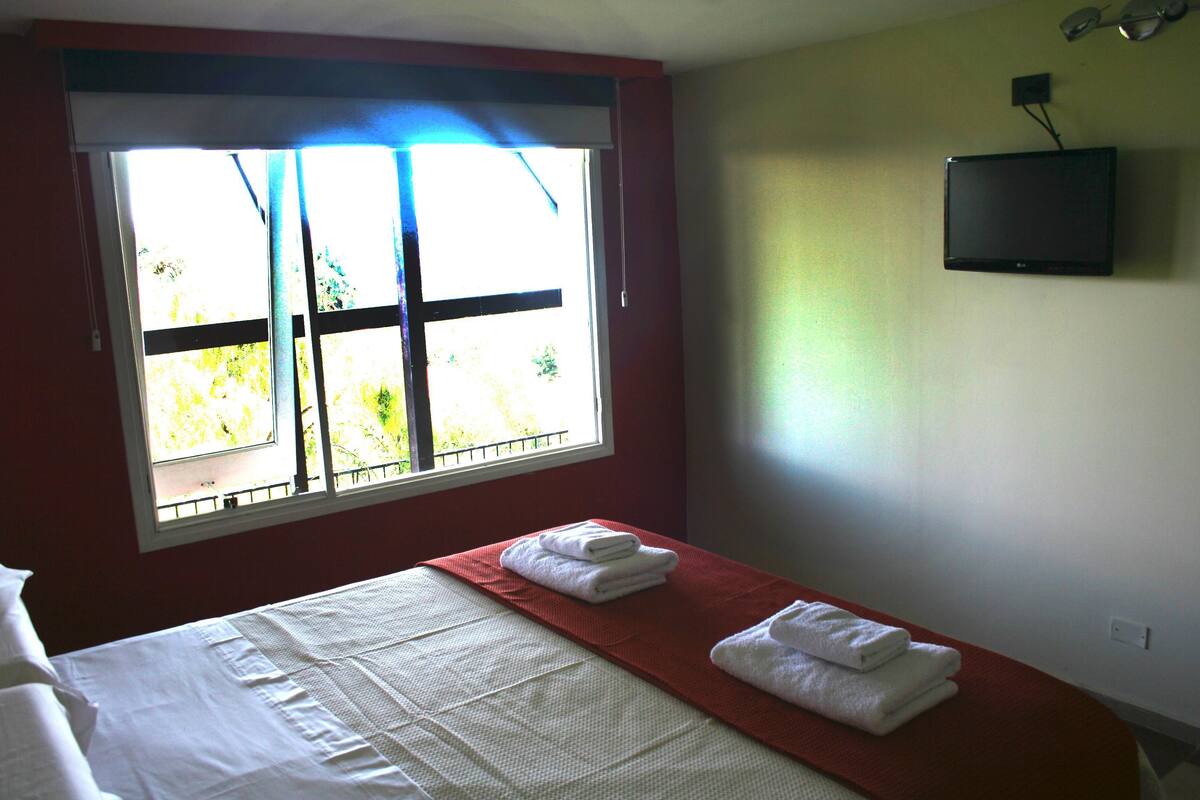 Quarto de hotel com uma cama, uma janela com persianas e uma televisão de tela plana fixada na parede.