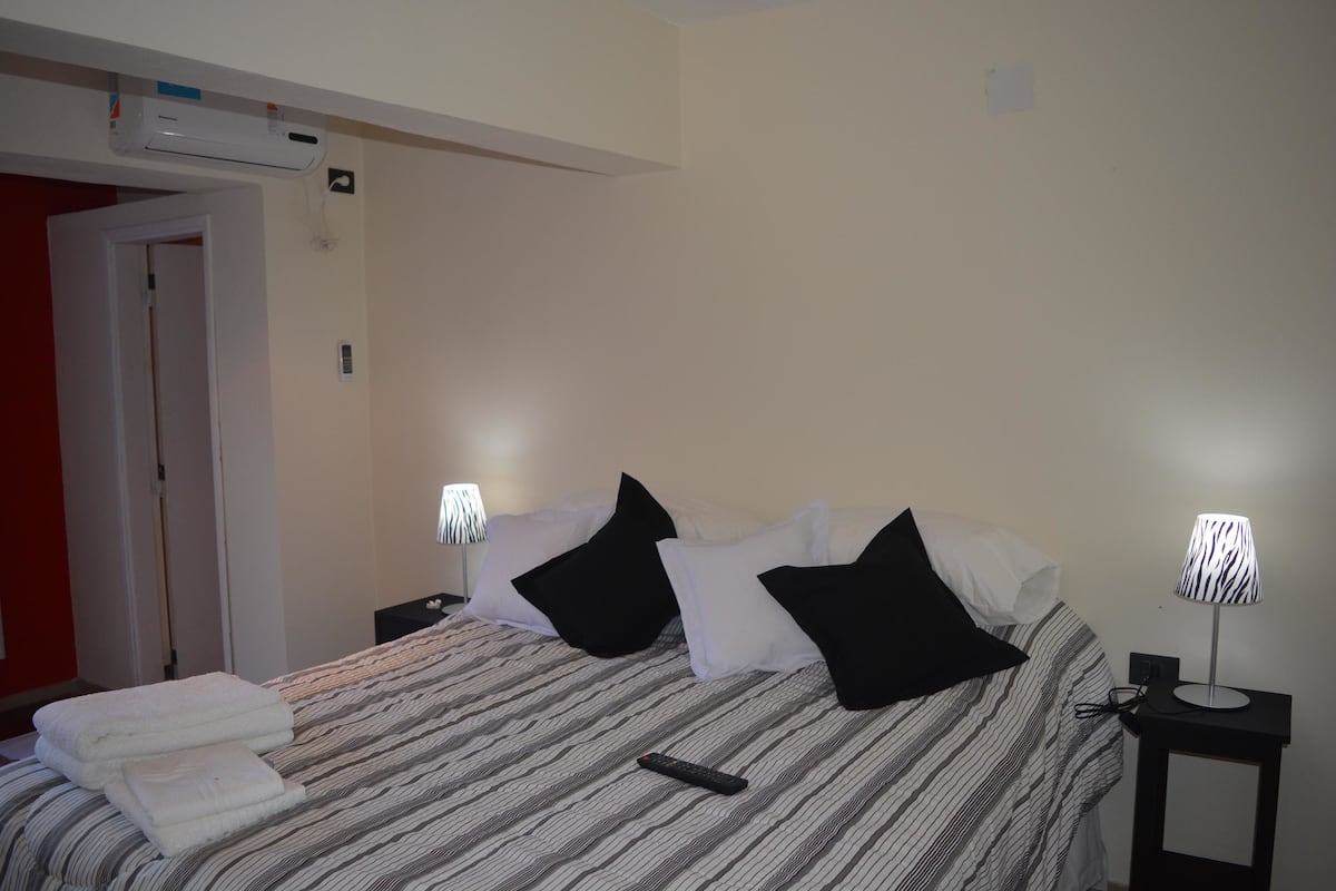 Quarto de hotel com cama, comando remoto, luminária e ar-condicionado fixado na parede.