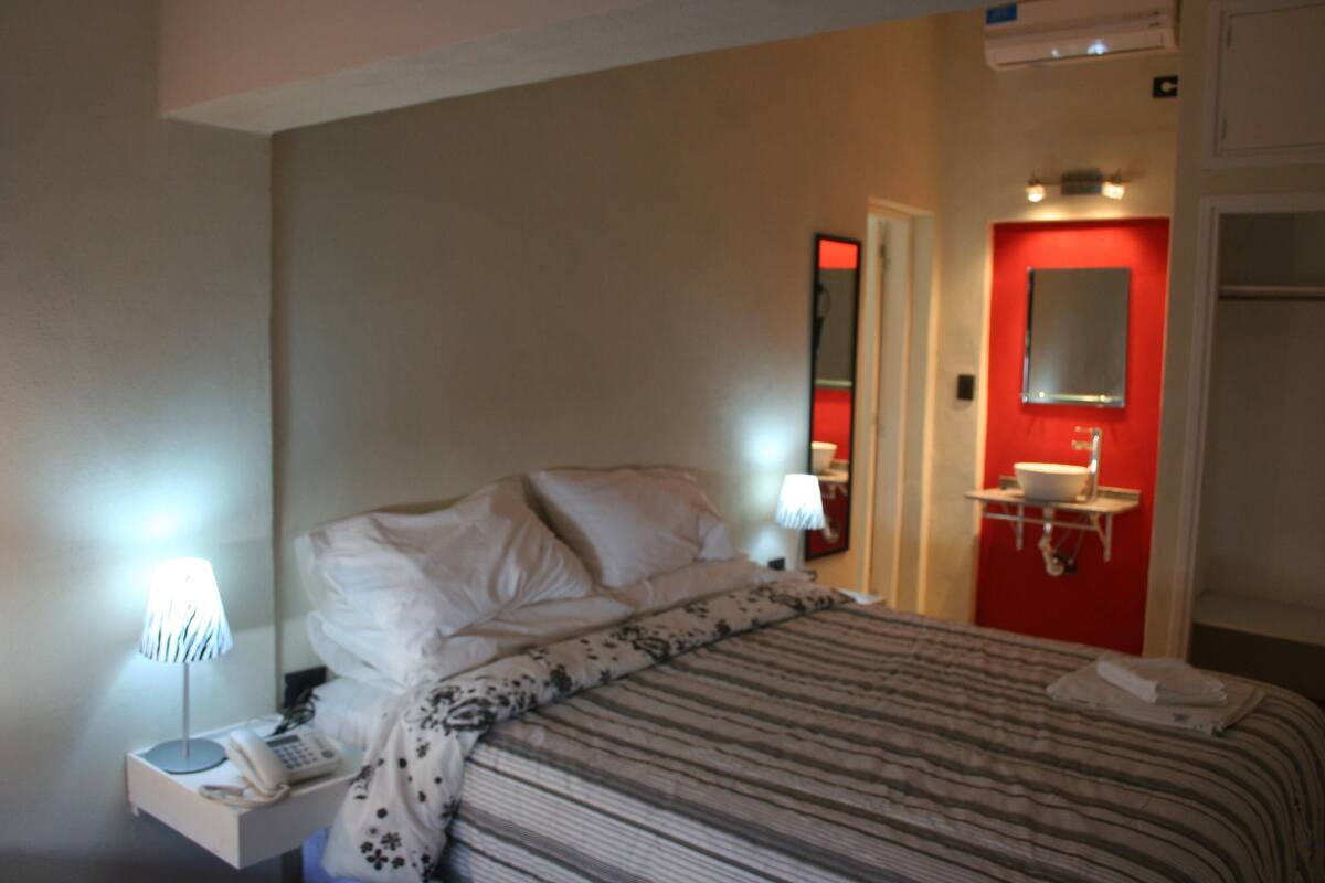 Quarto de hotel com cama, abajur, telefone e banheiro vermelho com espelho.