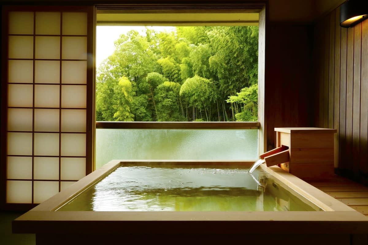 Un onsen japonés tradicional con una bañera de madera, vista a un bosque por una ventana y una puerta corredera.