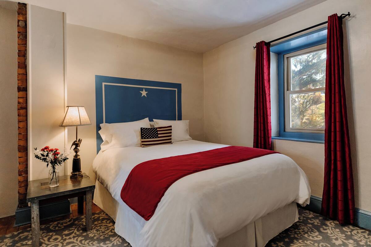 A neatly made bed with a red blanket, a small table with a lamp and flowers, and a window with red curtains.