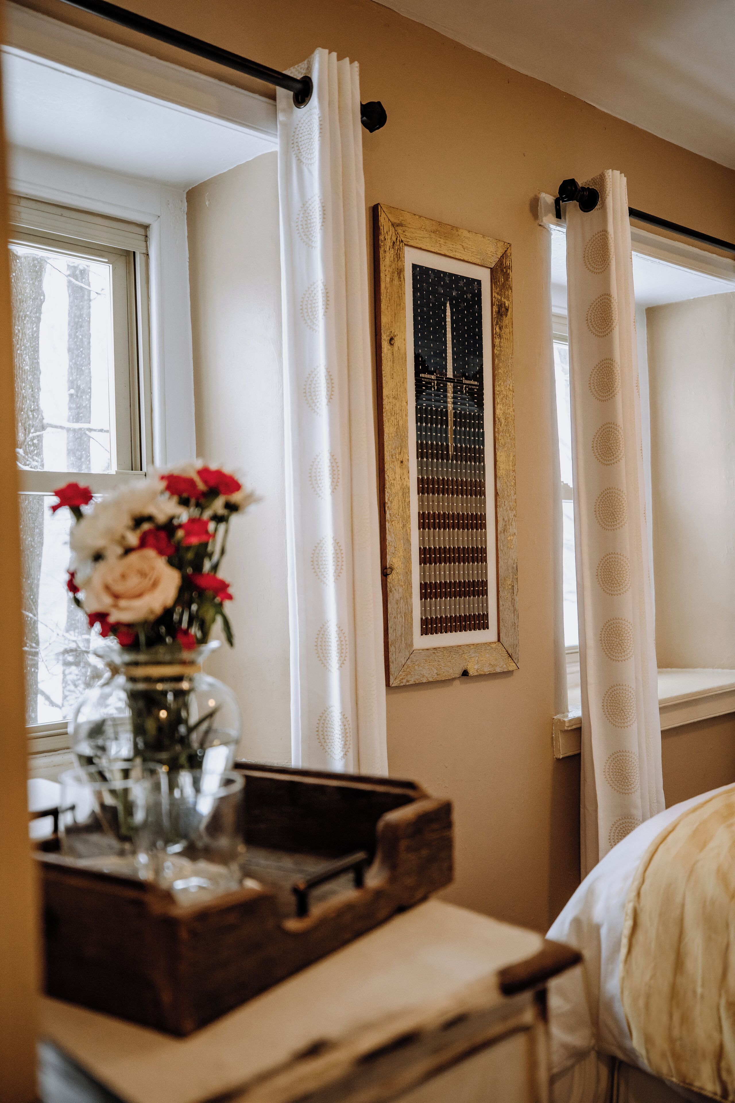 A room with a bed, a wooden nightstand, a vase with flowers, a framed picture, and a window with curtains.