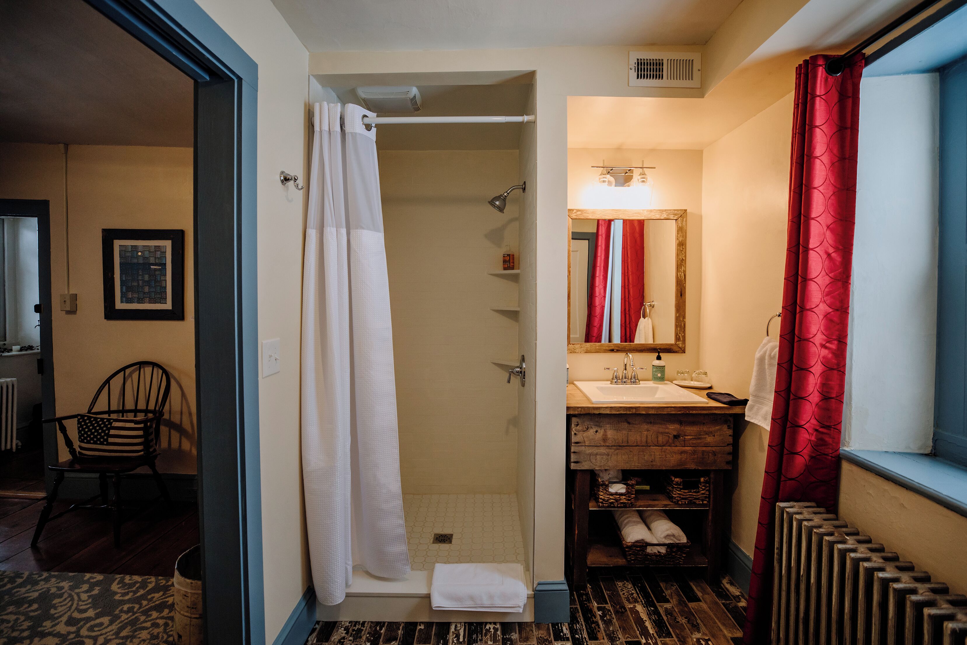 A bathroom with a shower, sink, and mirror. There is a chair and a radiator in the room.