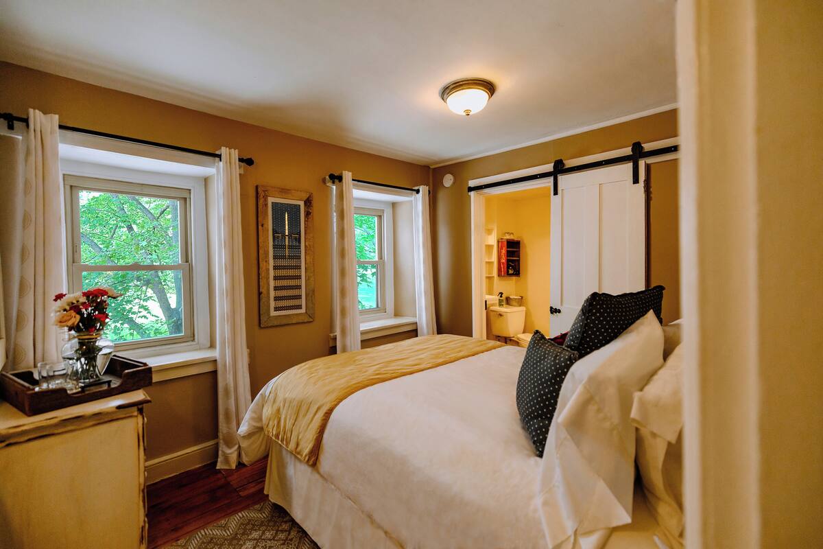 A bedroom with two windows, a bed with white bedding, a bedside table with a vase of flowers, and a nightstand.