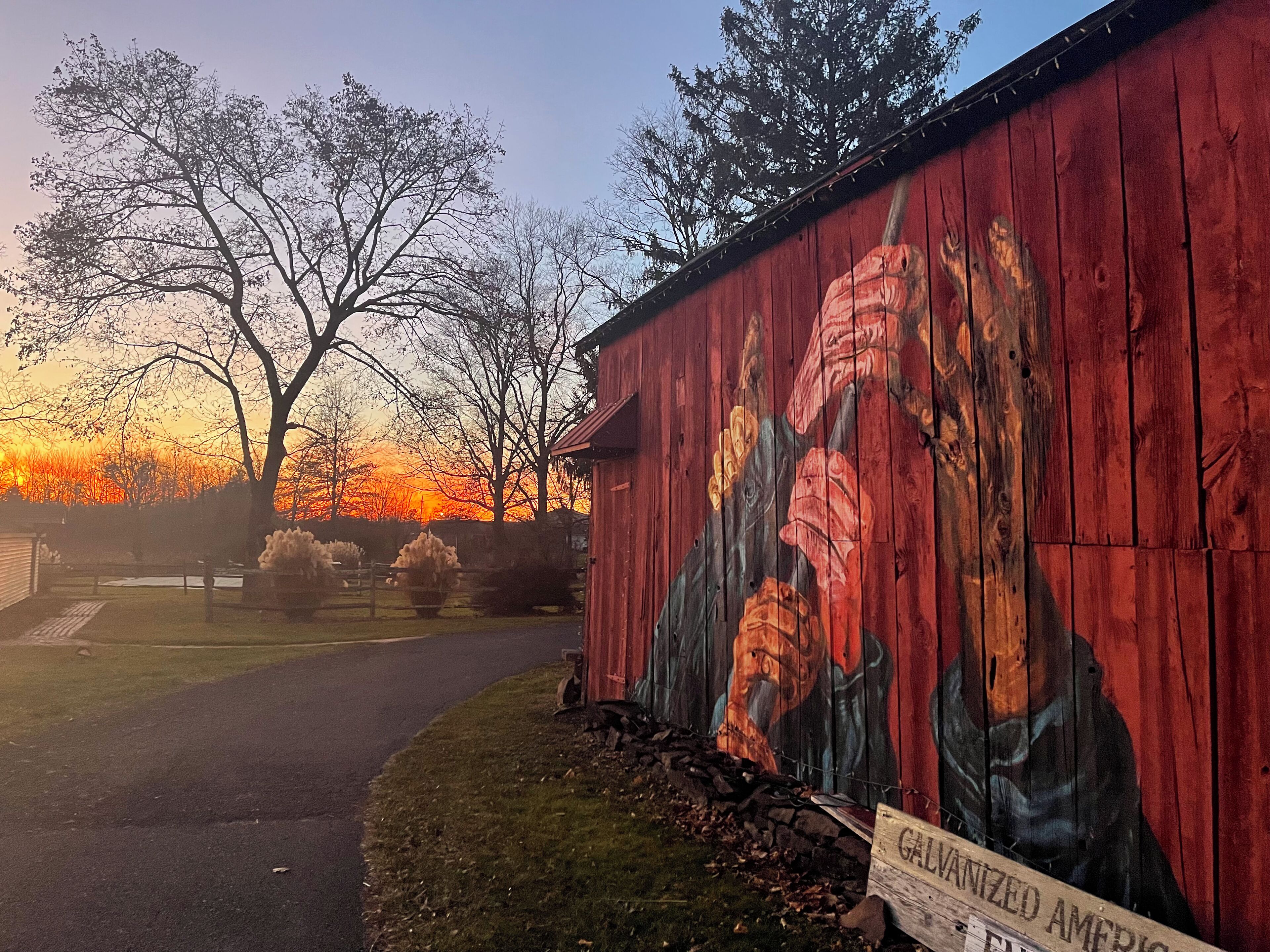 A red wooden building with a mural of a person holding flowers, a sign reading "GIVENAMED AMERICA," and a sunset in the background.