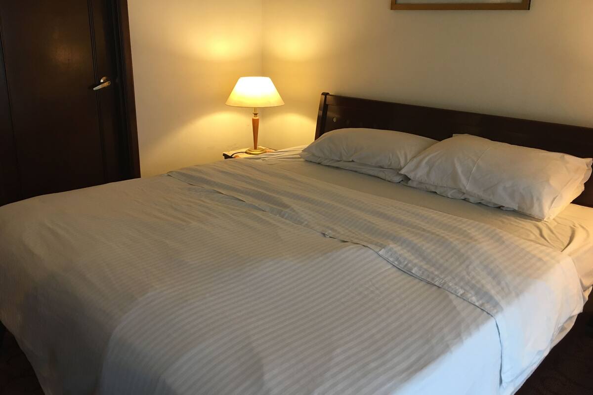 A neatly made bed with white linens and two pillows, a bedside lamp, and a framed picture on the wall.