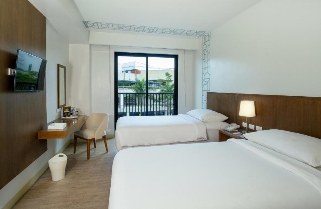 A hotel room with two beds, a desk, a chair, a TV, and a balcony with a view.