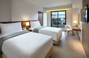 A hotel room with two beds, a desk with a chair, a TV, and a balcony with a view of buildings.