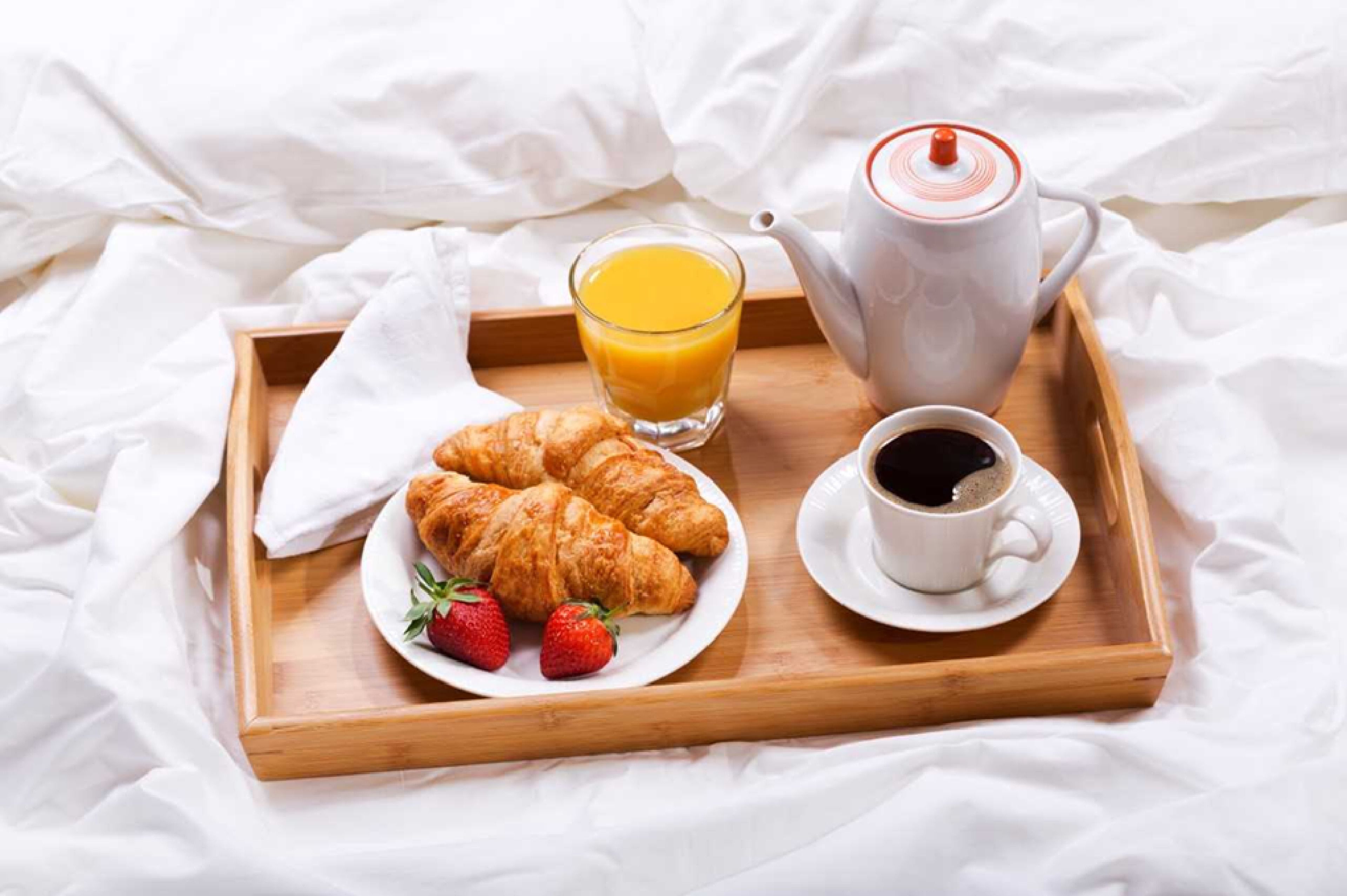 Una bandeja de madera con cruasanes, fresas, un vaso de jugo de naranja, una taza de café y una tetera sobre una cama con sábanas blancas.