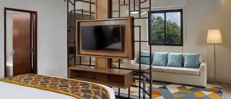 A hotel room with a bed, a TV on a wooden stand, a sofa with cushions, a lamp, and a window with a view of trees.