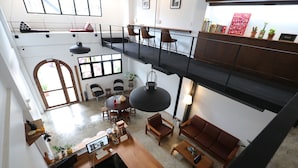 A spacious loft-style hotel room with a high ceiling, a mezzanine level, a dining area, and a seating area with a sofa and chairs.