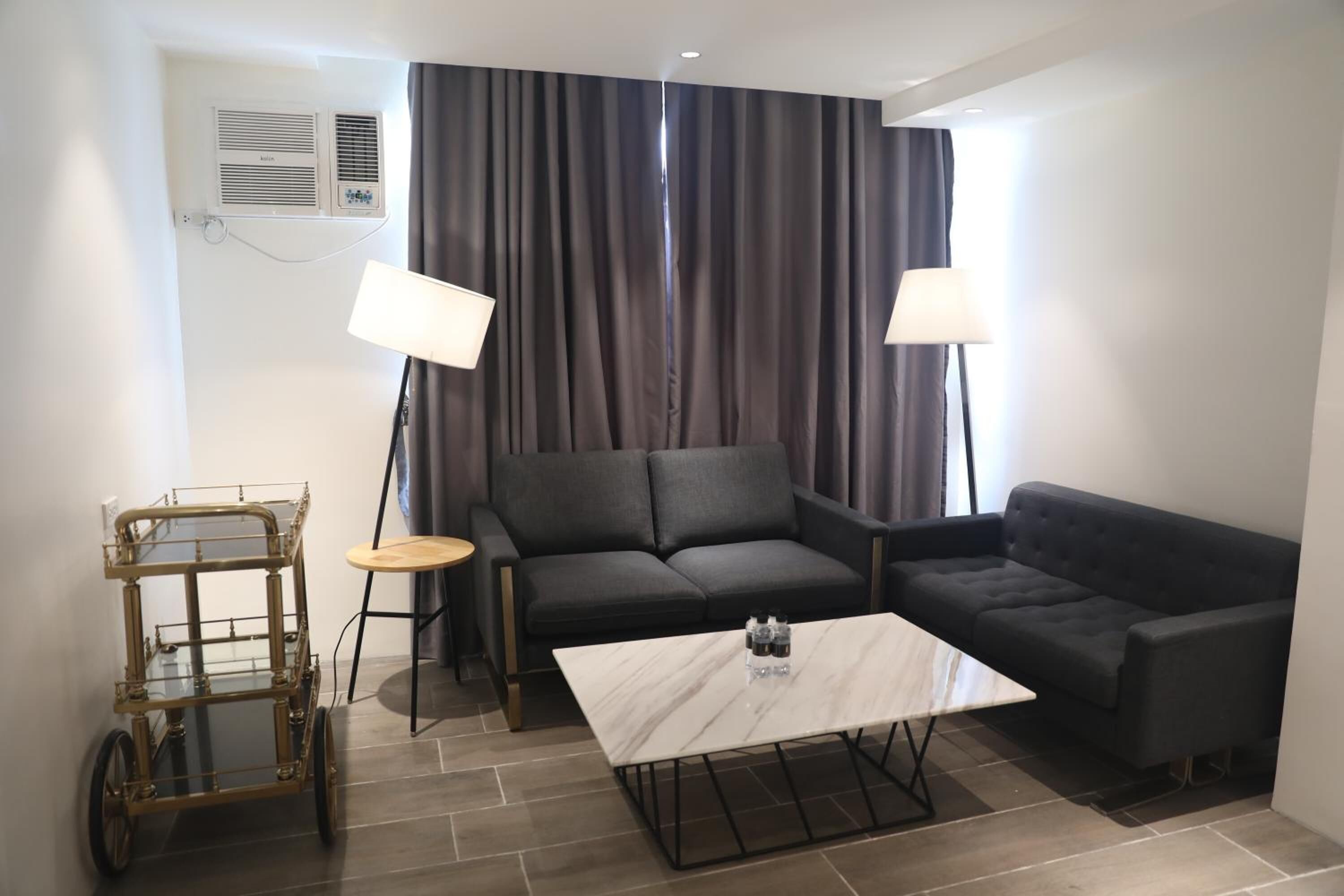 A modern living room with a dark grey sofa, a white marble coffee table, and a small side table. There is a gold-framed decorative cart and a wall-mounted air conditioning unit.