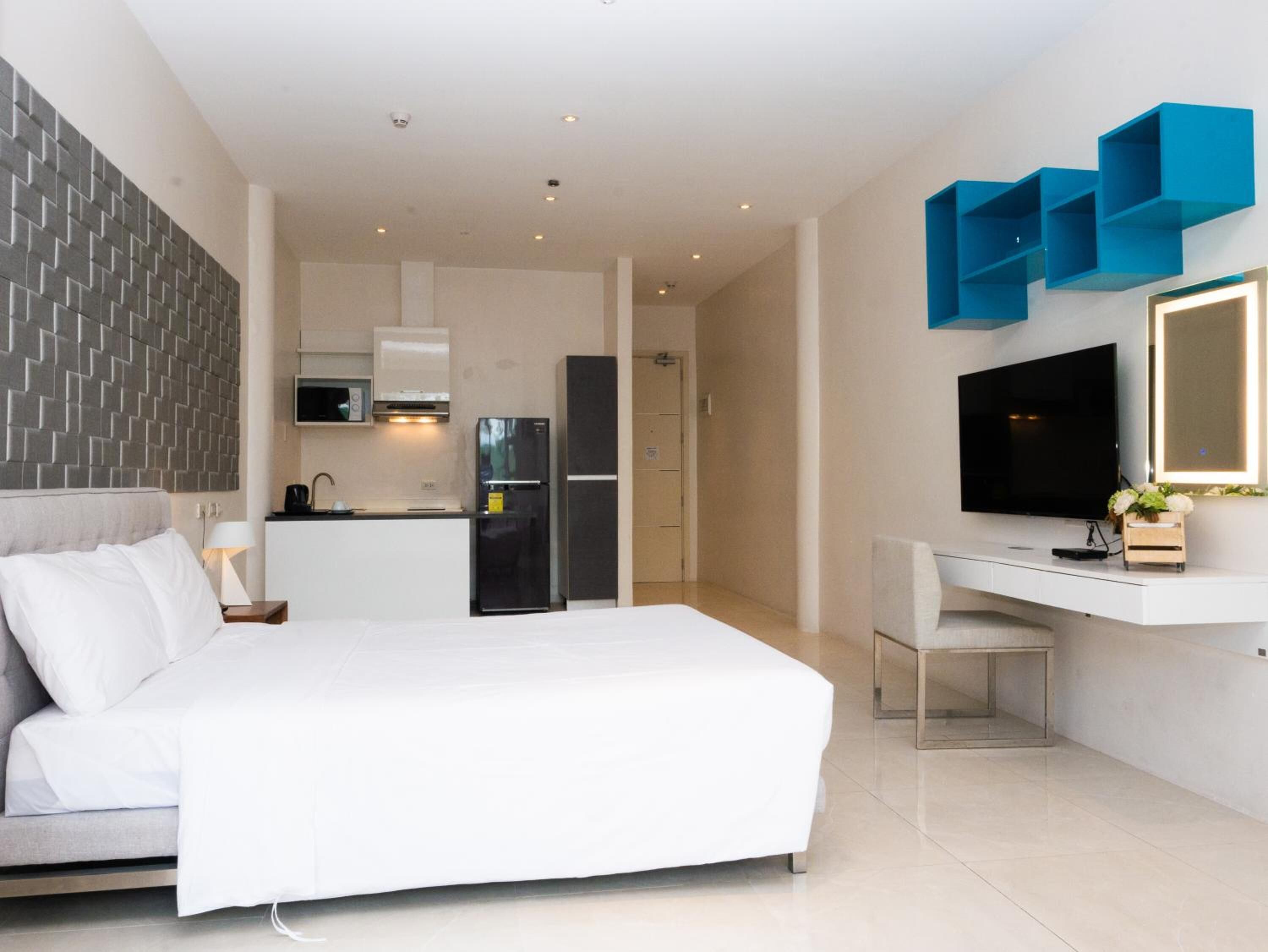 A modern hotel room with a bed, a desk, a TV, and a kitchenette.