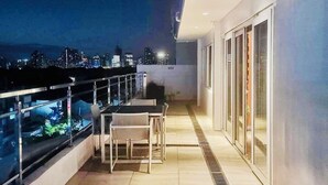 Een balkon met een tafel en stoelen, uitzicht op de stad 's nachts, en een glazen deur naar een binnenruimte.