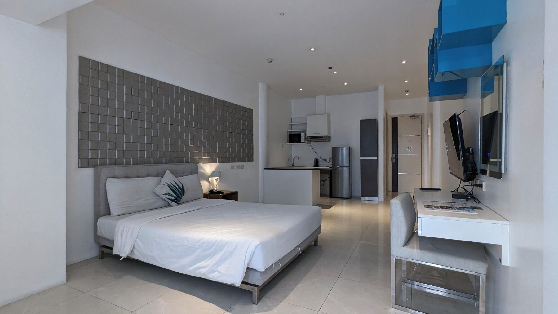 A modern hotel room with a bed, bedside table, lamp, and a desk with a computer.