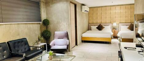 A modern hotel room with a sofa, armchair, glass coffee table, and a bed with a headboard.