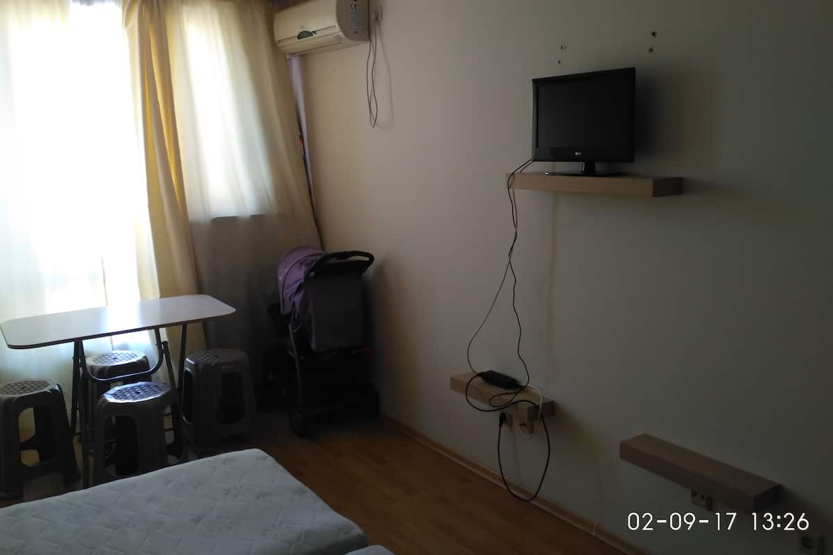 Une chambre d’hôtel comprenant un lit, une table, un téléviseur fixé sur une étagère en bois, une chaise, un petit tabouret, un appareil de climatisation et une fenêtre avec des rideaux.