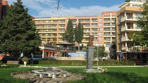 A courtyard with a fountain, benches, and a statue. In the background, there is a multi-story building with balconies and air conditioning units. - Vadim Apartments Vigo Panorama (Nessebar)