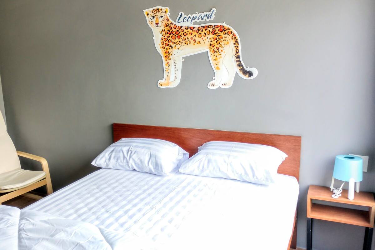 Ett hotellrum med en säng, en stol, en väggdekor med ett leopardmotiv och ett nattduksbord med en lampa.