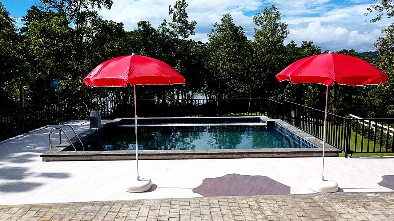 Una piscina con due ombrelloni rossi, circondata da un pavimento in mattoni e un recinto metallico.