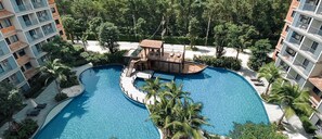 Une grande piscine incurvée, avec une structure en forme de bateau en bois au centre, entourée de palmiers et de bâtiments modernes.