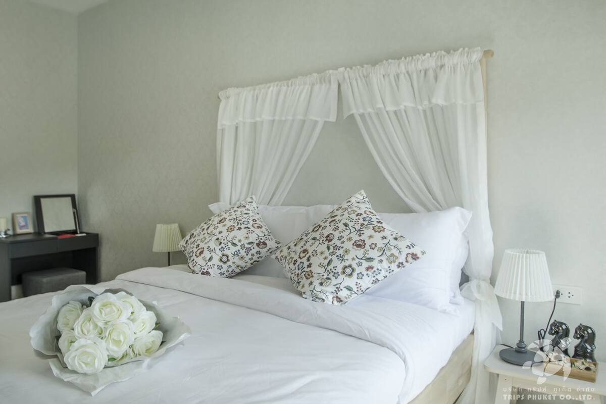 Ein ordentlich gemachtes Bett mit weißer Bettwäsche, Kissen mit Blumenmuster und einem darüberliegenden Baldachin.