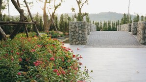 Uma trilha pavimentada conduz a um muro de pedra, rodeada por vegetação exuberante e plantas floridas.