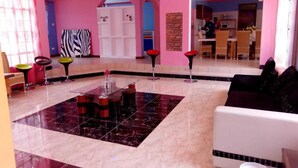 A spacious living area with a central coffee table, a sofa, and a dining table with chairs. There is a kitchen area in the background with a person visible. - The Joshua Gardens (Nairobi)