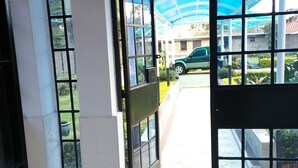 An open door leading to a covered outdoor area with a tiled floor and a view of a parked car and greenery outside. - The Joshua Gardens (Nairobi)