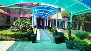 A covered walkway leading to a building with a pink facade and a blue roof. - The Joshua Gardens (Nairobi)
