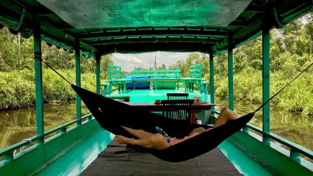 A hammock tied between two poles on a boat with a table and chairs in the background. - Houseboat Bee Kelotok (Kumai)
