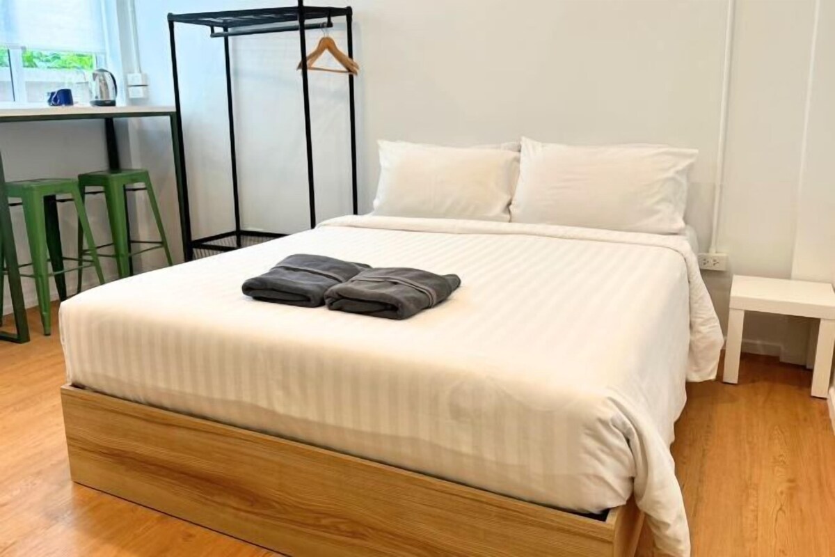 Una cama bien tendida con sábanas blancas y una manta gris doblada, un cabecero de madera, una mesita de noche pequeña y un taburete verde en una habitación luminosa con una ventana.