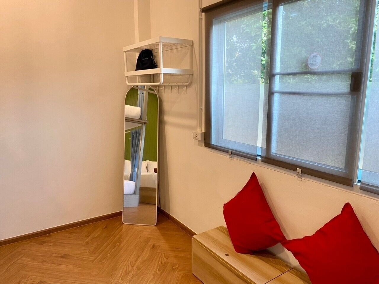 A room with a wooden floor, a window with blinds, a mirror, and a small white shelf with a black bag on it.
