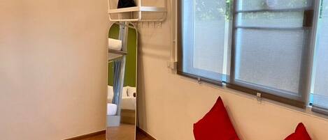 A room with a wooden floor, a window with blinds, a mirror, and a small white shelf with a black bag on it.