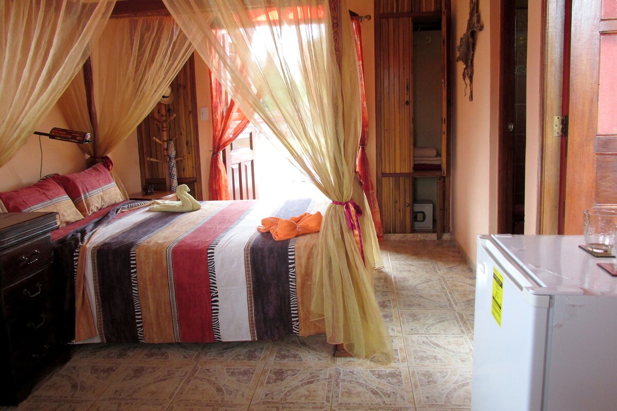 A bedroom with a bed, curtains, a nightstand, a dresser, and a refrigerator.