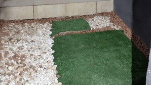 Un pequeño espacio al aire libre con una escalera de concreto que lleva a una puerta con un portón metálico. Hay una planta en maceta en los escalones y una sección de césped artificial.