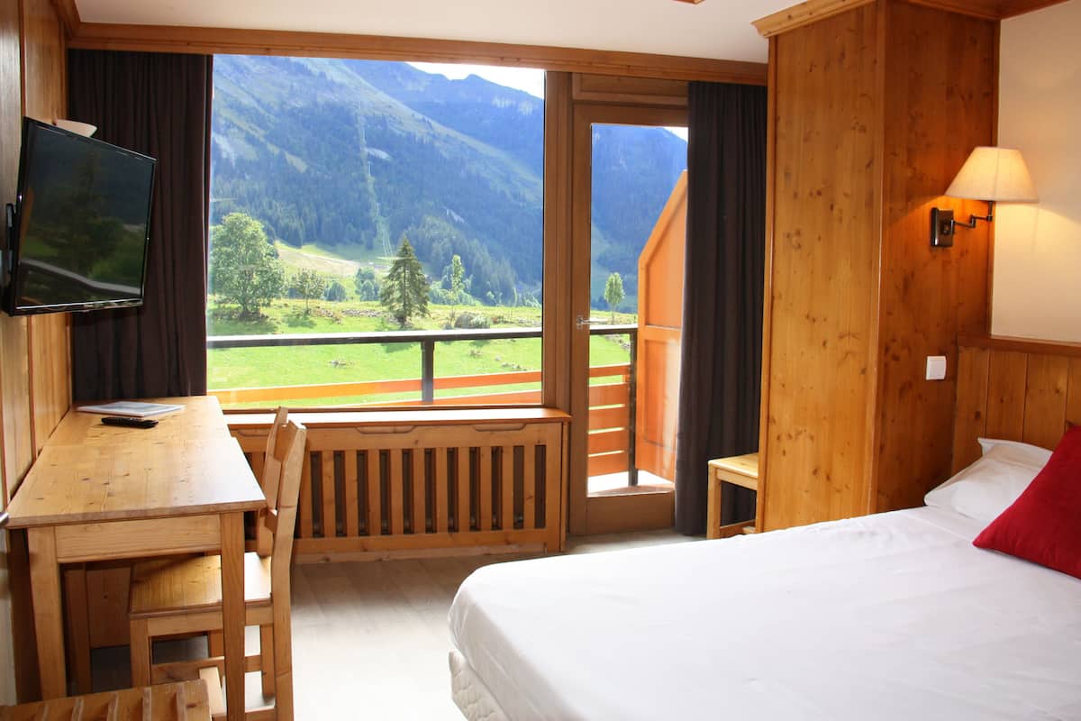 Habitación de hotel con un escritorio de madera, una cama con ropa de cama blanca, un almohilarrojo y vistas a montañas a través de un ventanal grande.